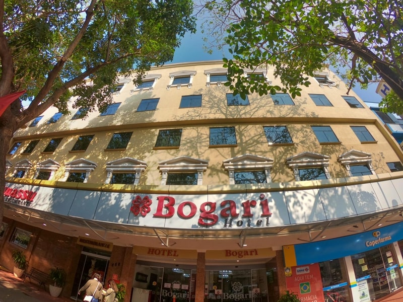 Fachada do Bogari Hotel, com arquitetura elegante em tons bege e branco. A entrada do hotel é ampla e convidativa, situada em uma rua arborizada e próxima de lojas e restaurantes, proporcionando comodidade aos hóspedes. As janelas grandes e simétricas dão um charme especial à fachada, e o letreiro vermelho destaca o nome do hotel. Ideal para turistas que procuram conforto, localização estratégica e fácil acesso às atrações de Foz do Iguaçu.
